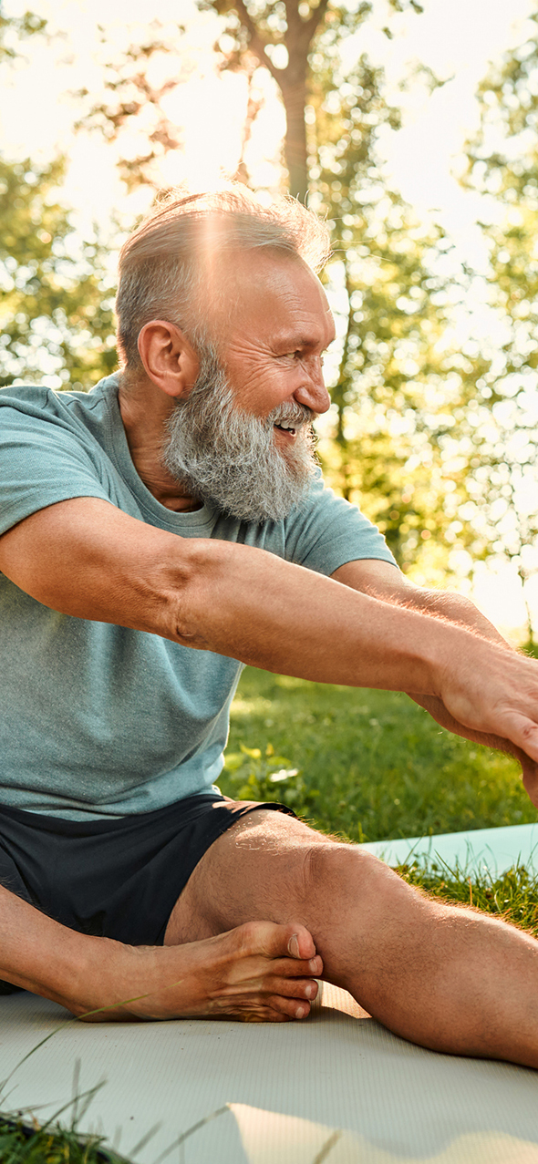 Sporty man with grey hair stretching on yoga mats with hands to one leg during outdoors workout.